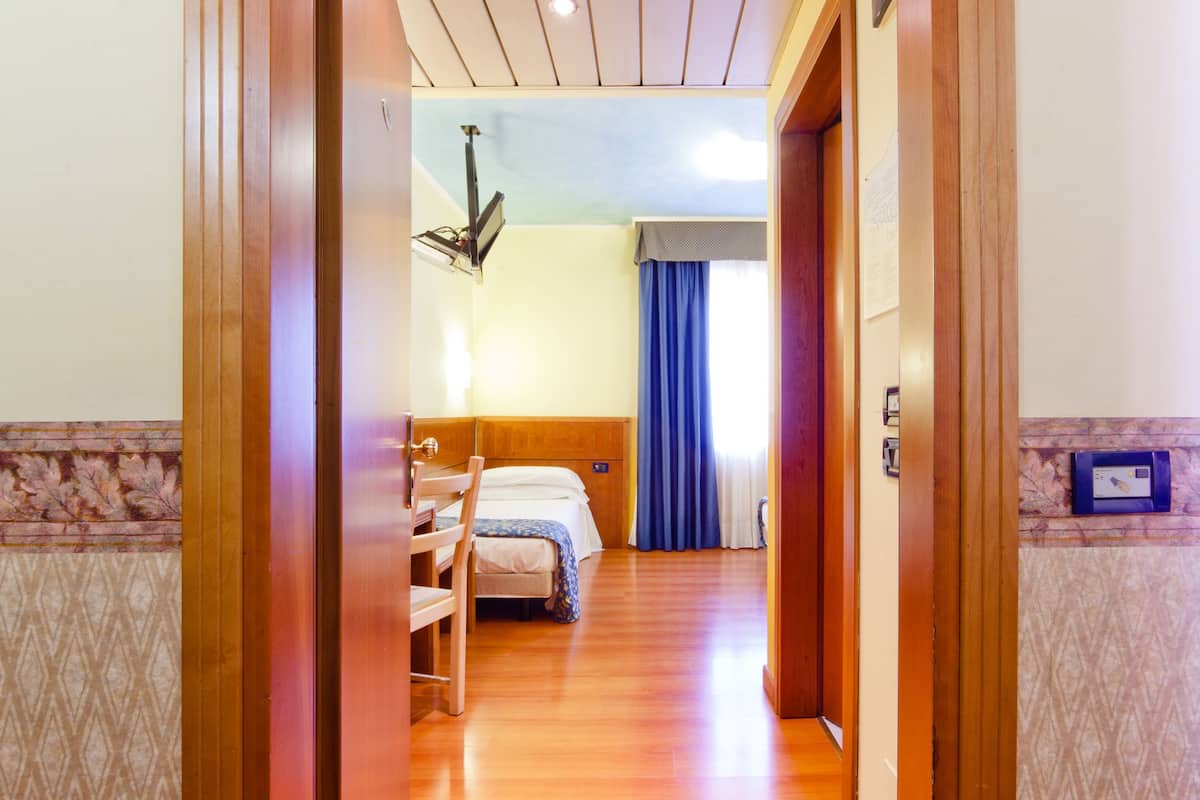 A hotel room with a wooden door, a bed with a blue bedspread, a desk, and a chair.