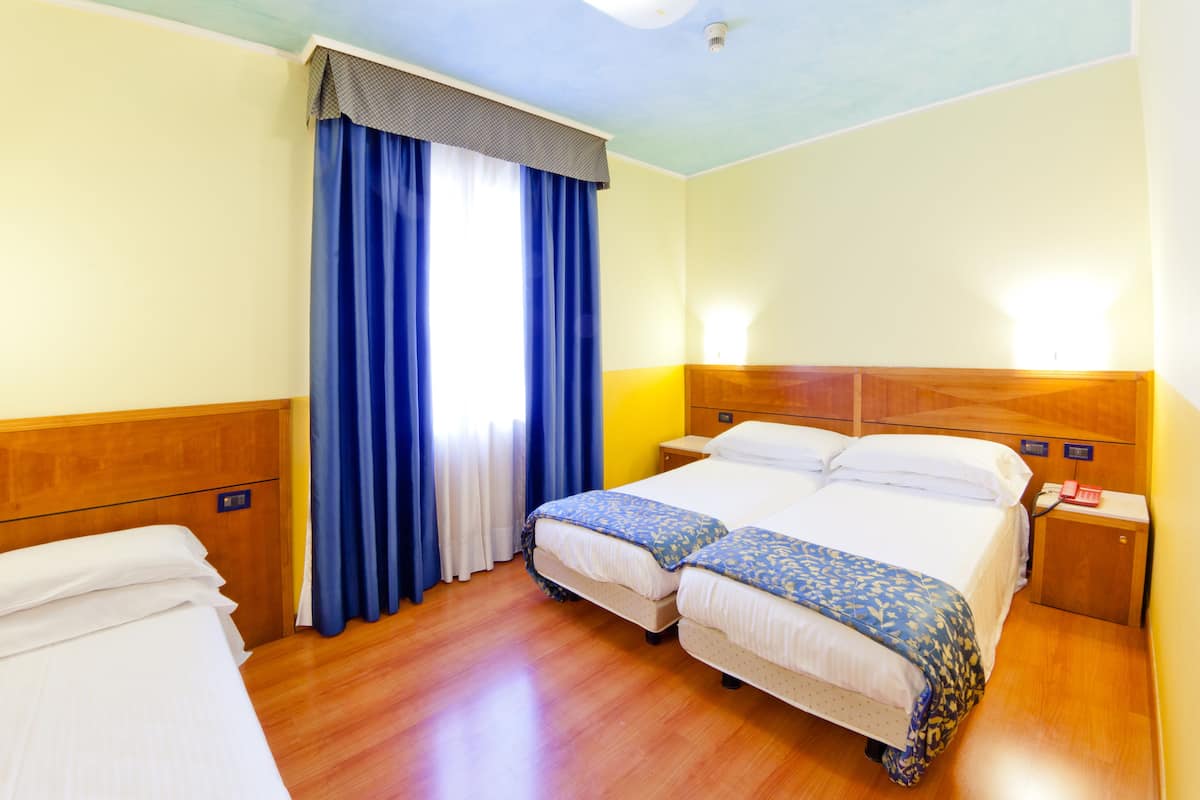 A hotel room with two beds, wooden headboards, a nightstand with a lamp, and a window with blue curtains.