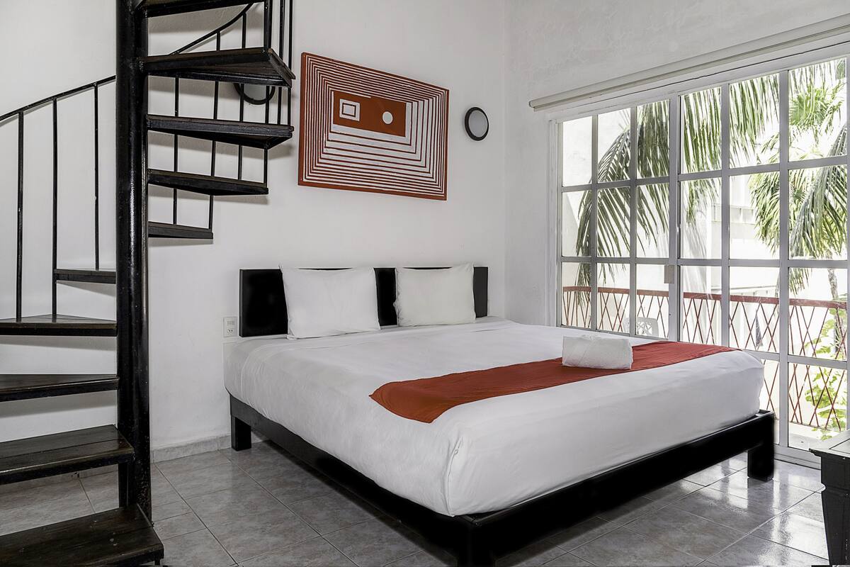 A bedroom with a bed, a wooden staircase, a wall clock, and a window with a view of palm trees.