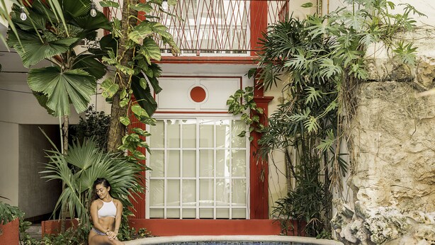 A woman sitting by a pool surrounded by lush tropical plants and a red and white building. - Hotel Maya Turquesa - Playa del Carmen (Playa del Carmen)