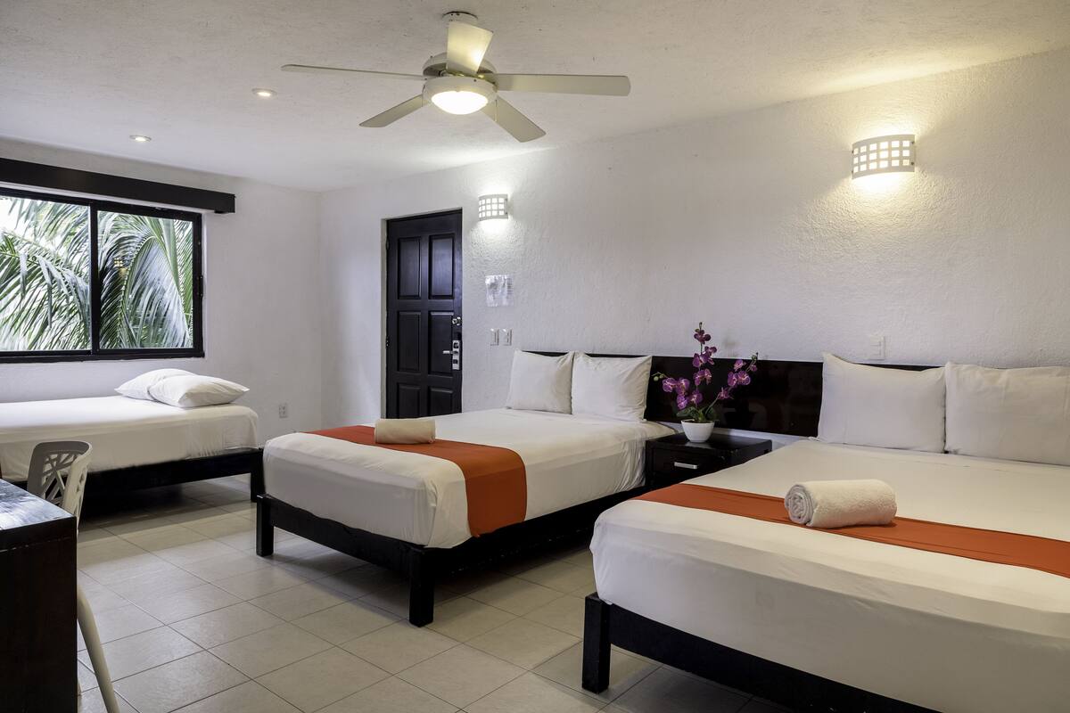 A hotel room with two beds, a desk, a chair, a ceiling fan, and a window with a view of palm trees.