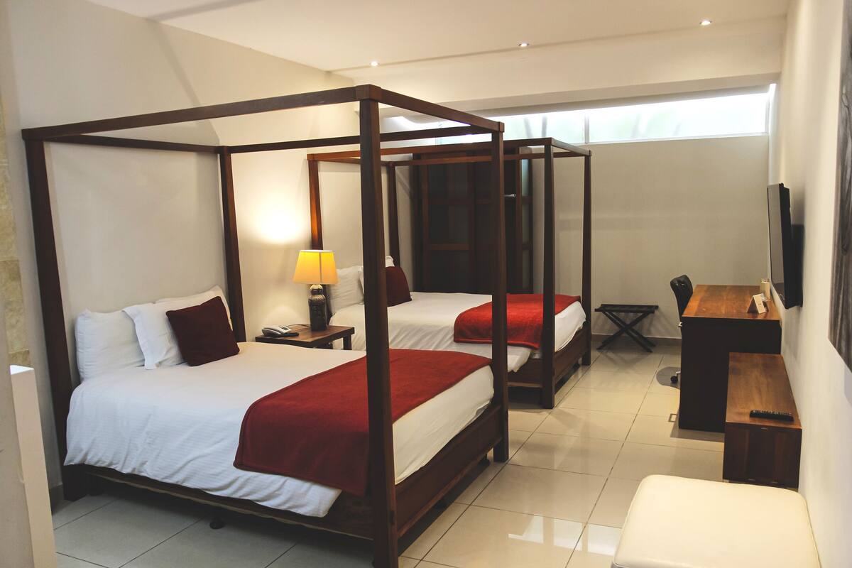 A four-poster bed with white linens and red accents, a desk with a chair, and a television on the wall.