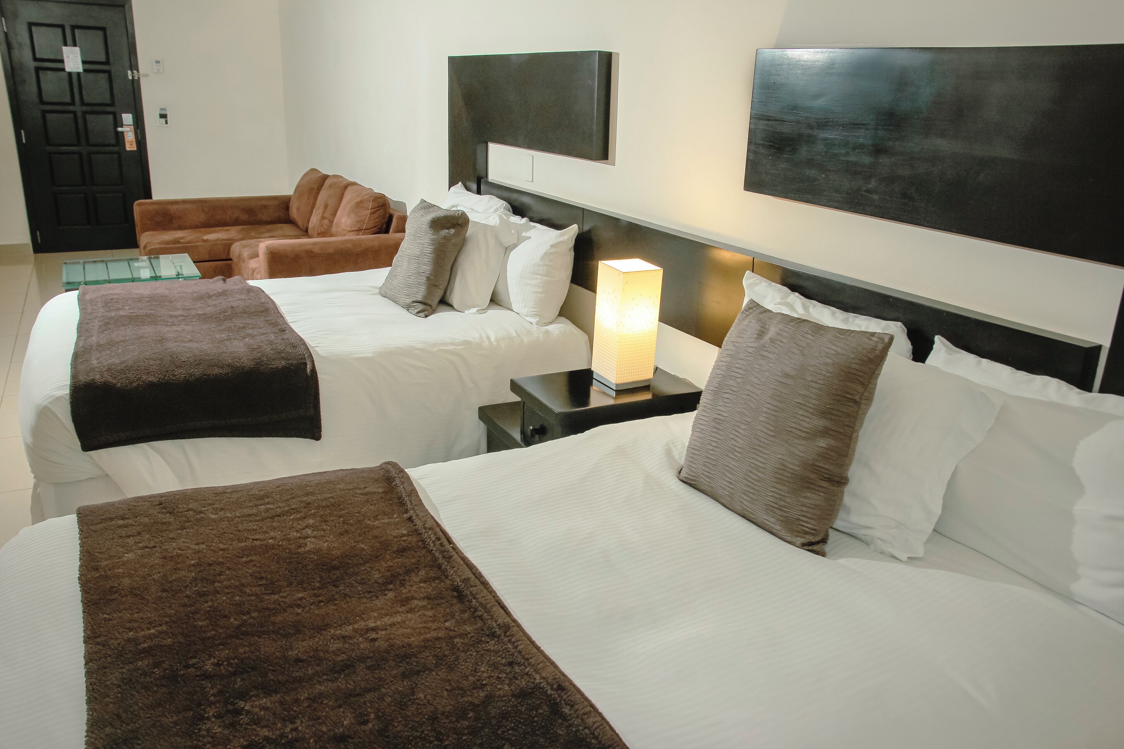 A hotel room with two beds, a nightstand with a lamp, a brown leather sofa, and a black door.