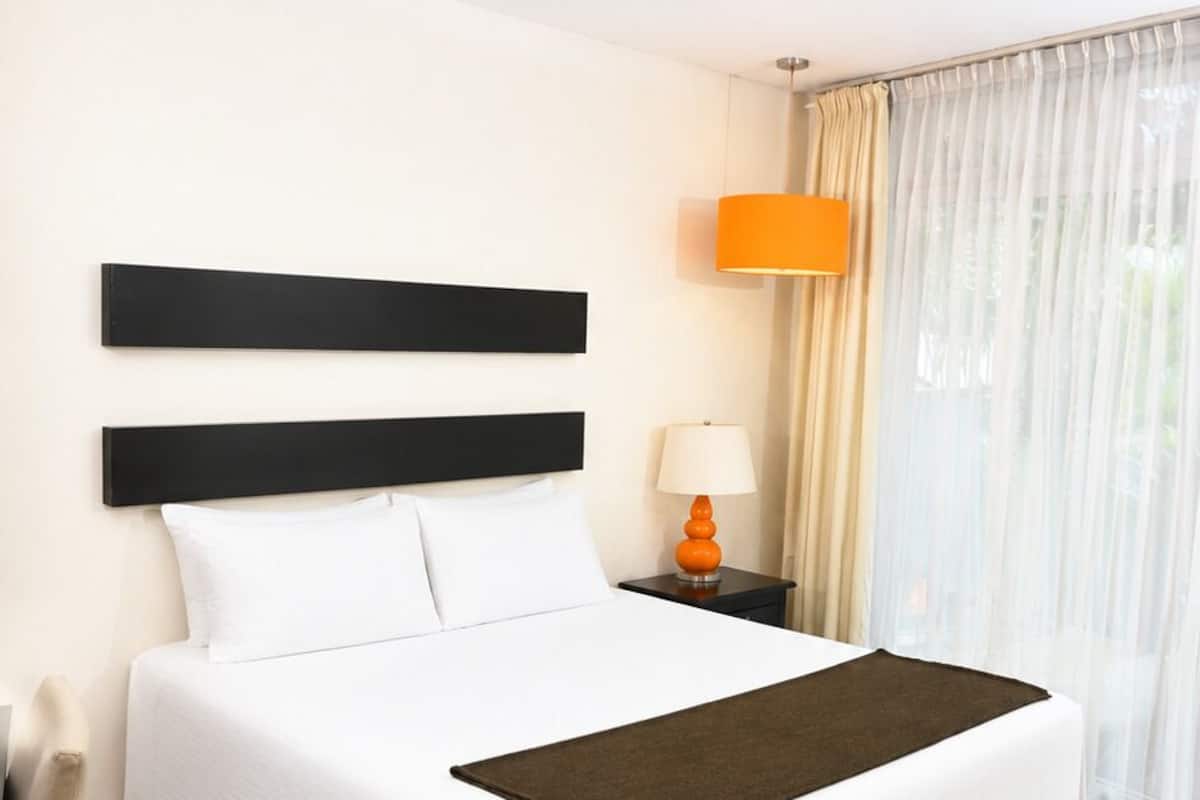 A hotel room with a large bed, a nightstand with a lamp, a chair, and a view of the outdoors through a window.