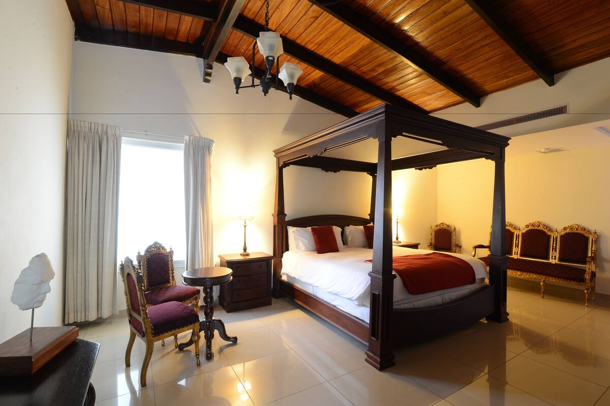 A four-poster bed with a wooden canopy, a sitting area with a chair and a small table, and a large window with curtains.