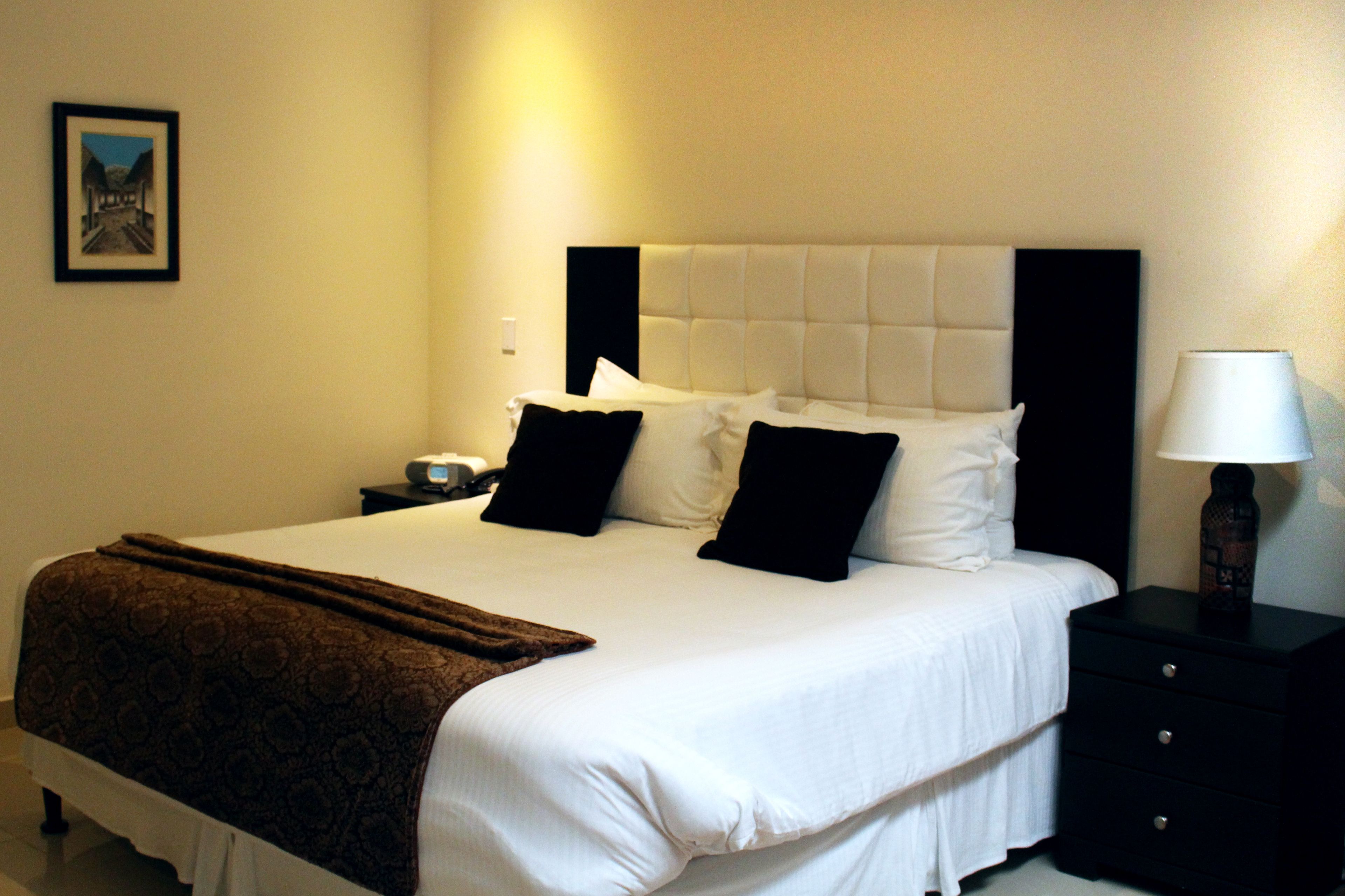 A neatly made bed with a brown blanket, a black nightstand with a lamp, and a framed picture on the wall.
