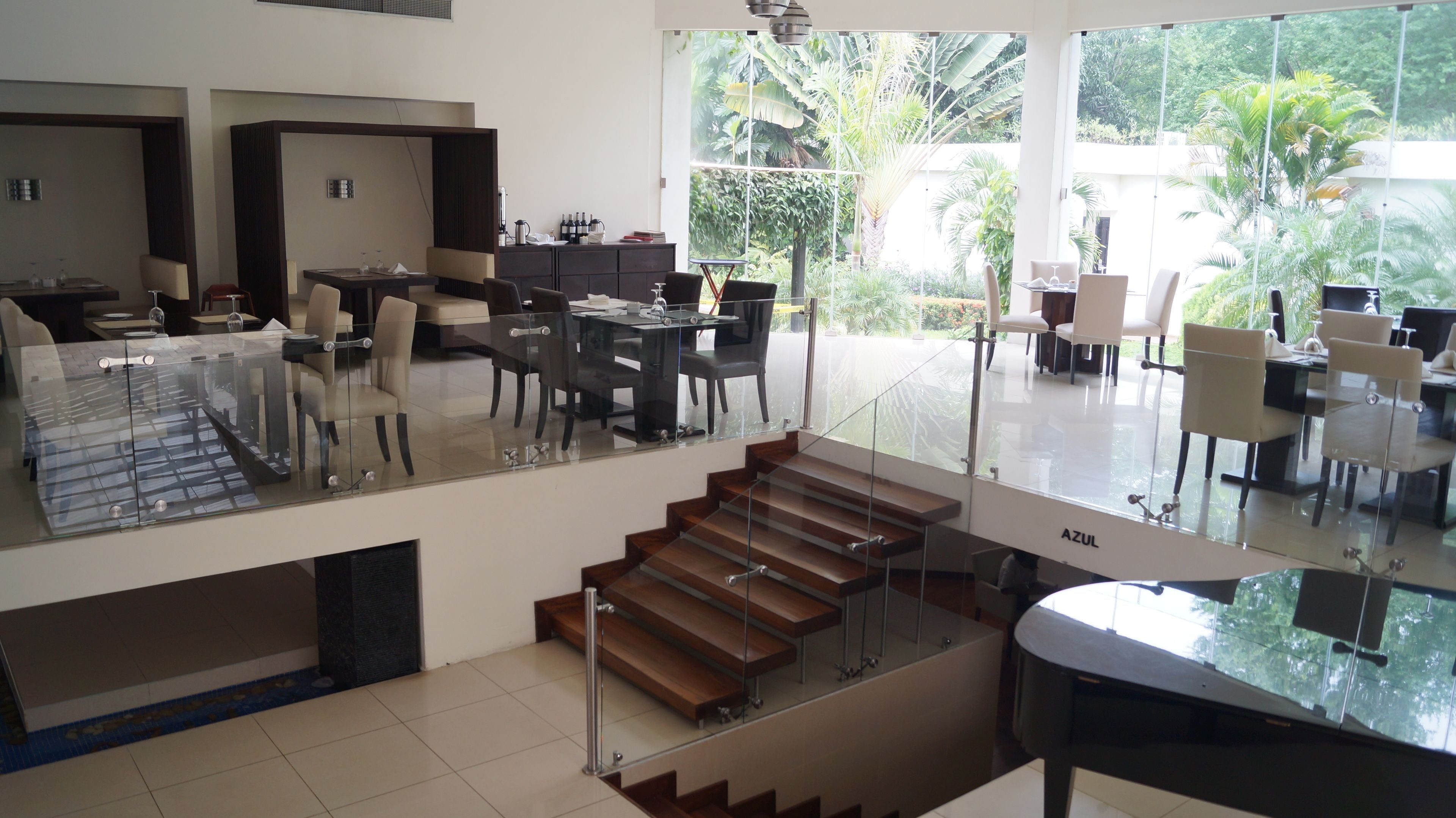 Un interior moderno con una escalera de cristal que conecta dos plantas, un comedor con mesas y sillas, y vistas a abundante vegetación exterior.