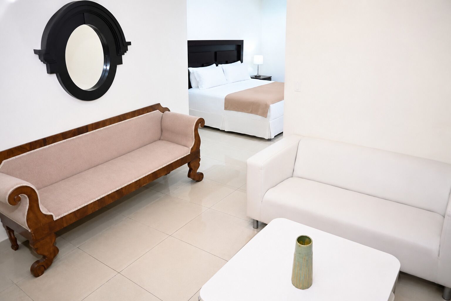 A room with a wooden bench, a white sofa, a small table with a vase, and a bed in the background.