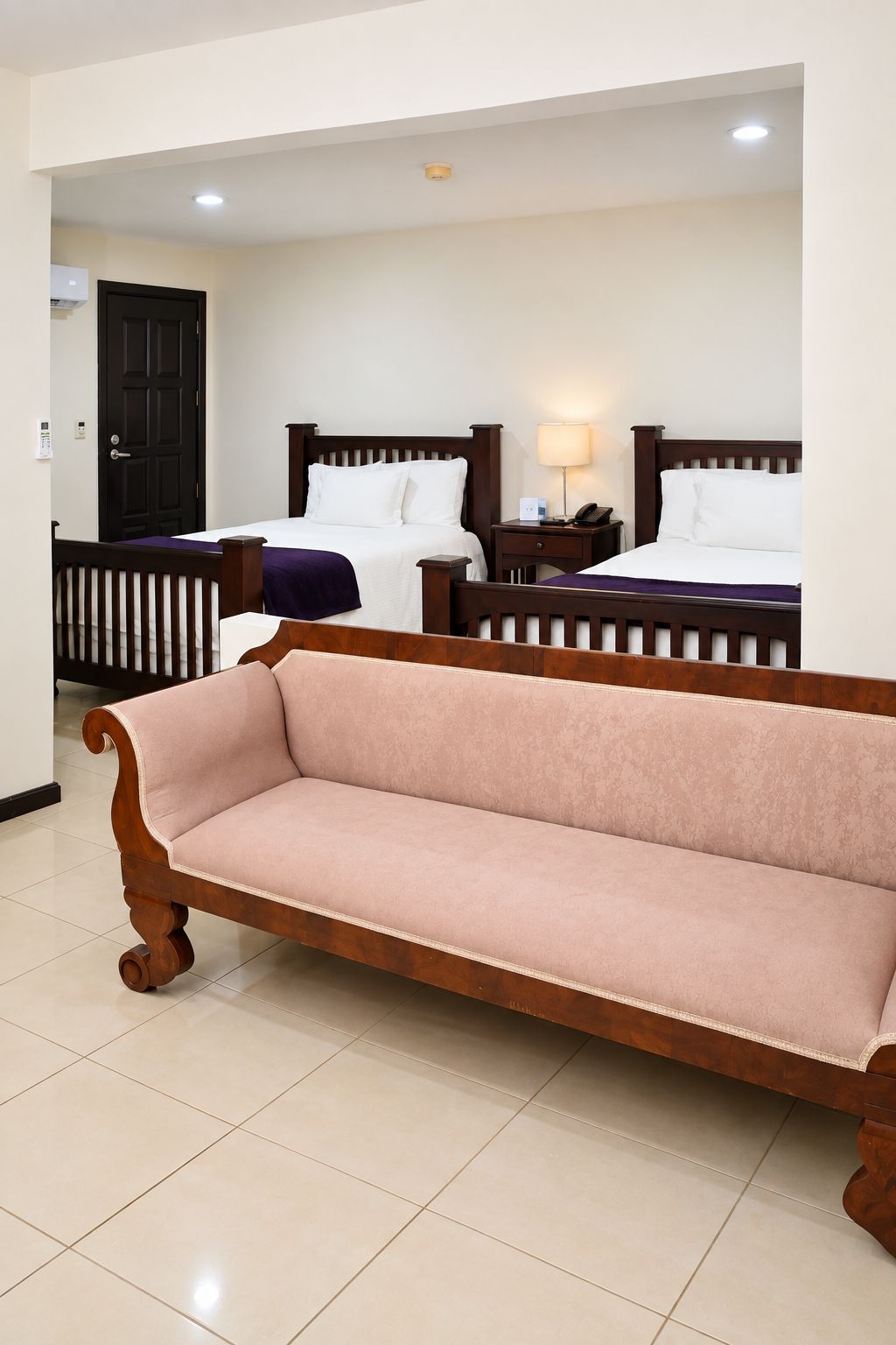A hotel room with two beds, a sofa, a door, and a wall-mounted air conditioning unit.