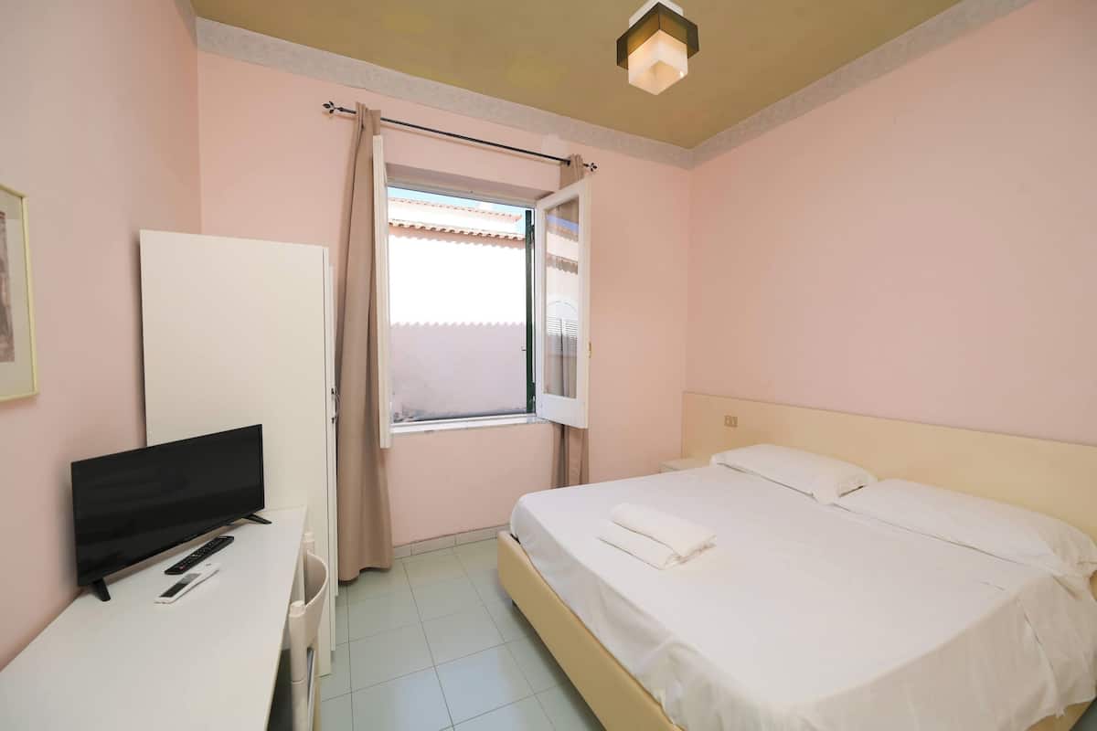 A hotel room with a bed, a desk with a TV and remote, a window with curtains, and a ceiling light.
