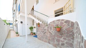 A narrow alleyway with a stone wall and a white building with a balcony. There are potted plants and a blue mat on the ground. - Hotel Villa Diana (Ischia)