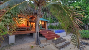 A tropical resort with a wooden deck, a pool with blue tiles, and a staircase leading to a covered area.