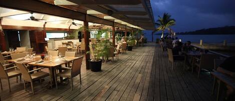 Un área de comedor al aire libre con mesas y sillas de madera, plantas en macetas y vista al agua.
