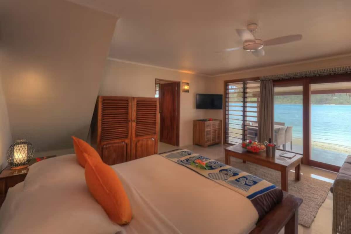 Een slaapkamer met een bed, een televisie, een raam met gordijnen en uitzicht op het water.