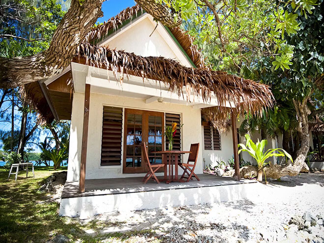 Een strandhuis met rieten dak, grote ramen en een eethoek met stoelen.