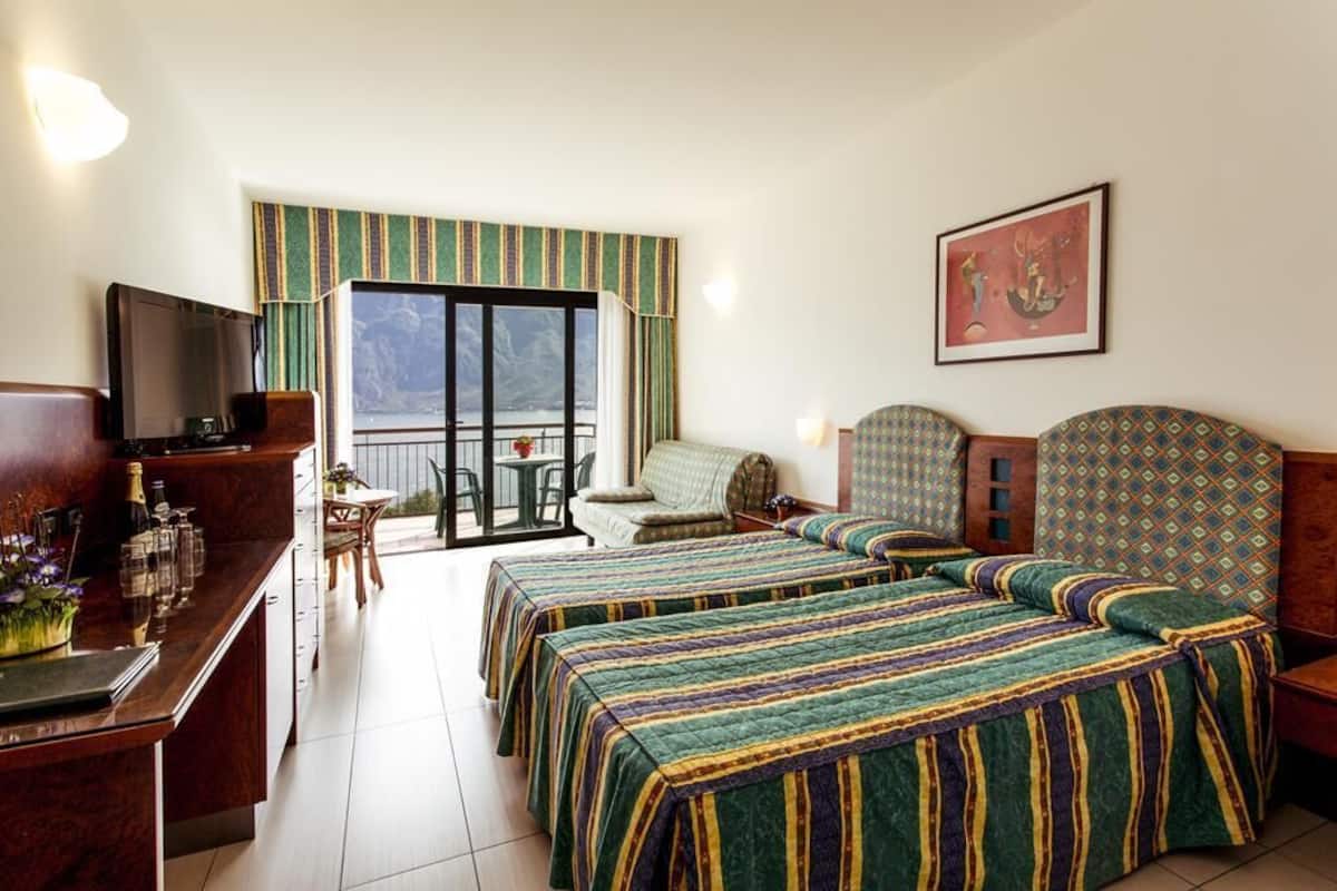 A hotel room with two beds, a TV, a desk with a computer, a balcony with a table and chairs, and a painting on the wall.