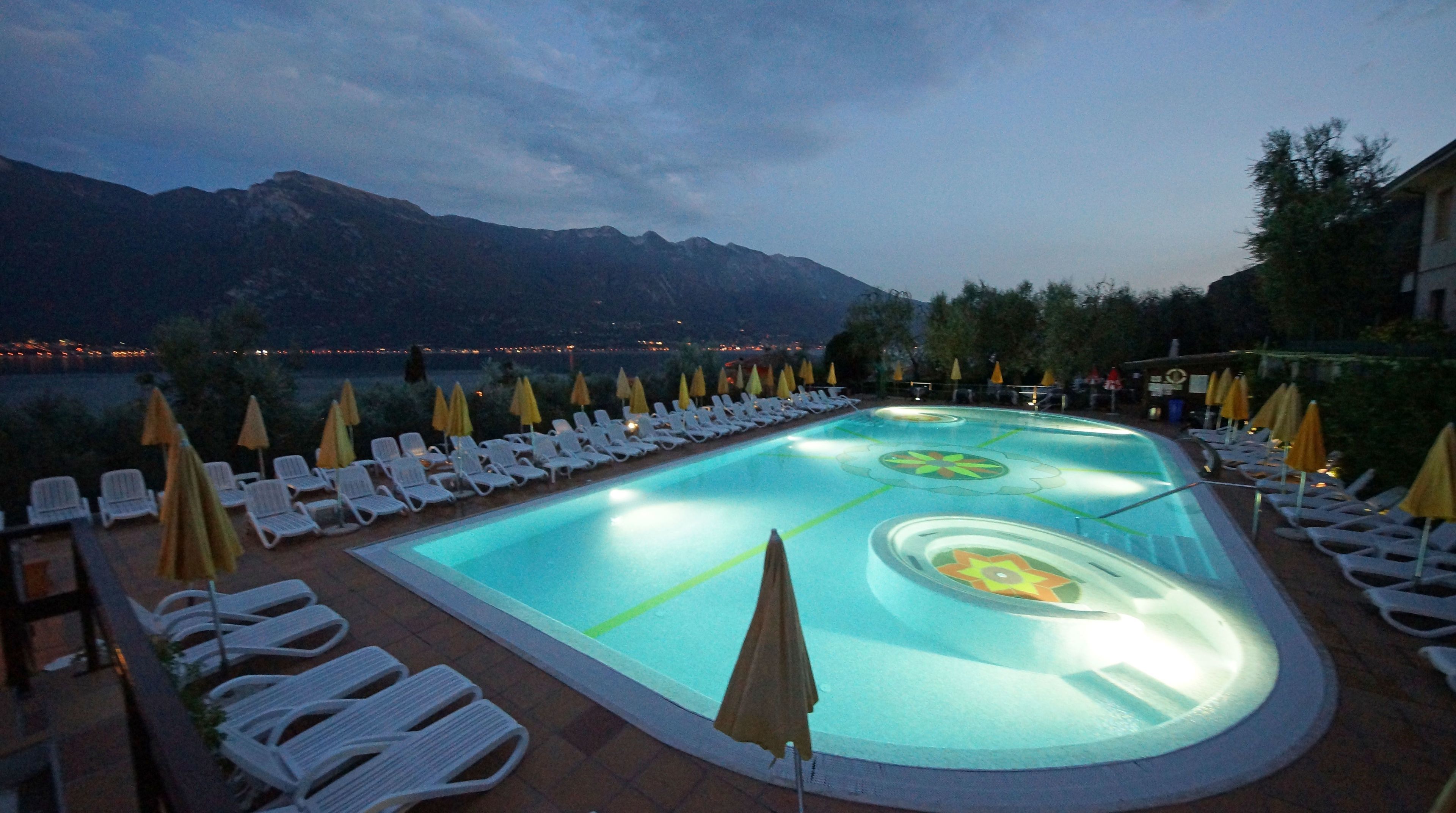 A swimming pool with lounge chairs and umbrellas, surrounded by mountains and a view of a lake.