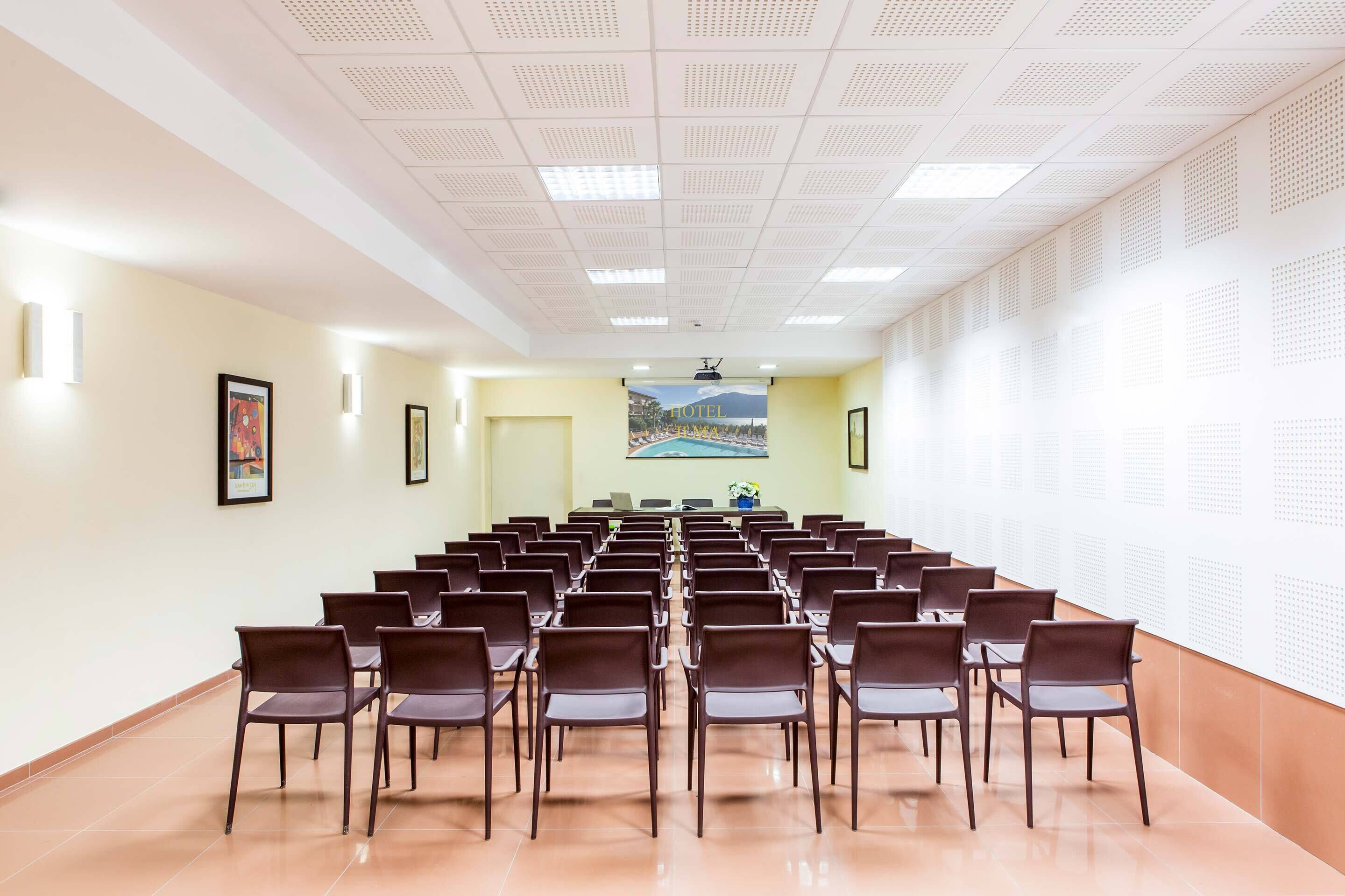 A conference room with rows of chairs facing a presentation screen, a large poster on the wall, and a door at the far end.