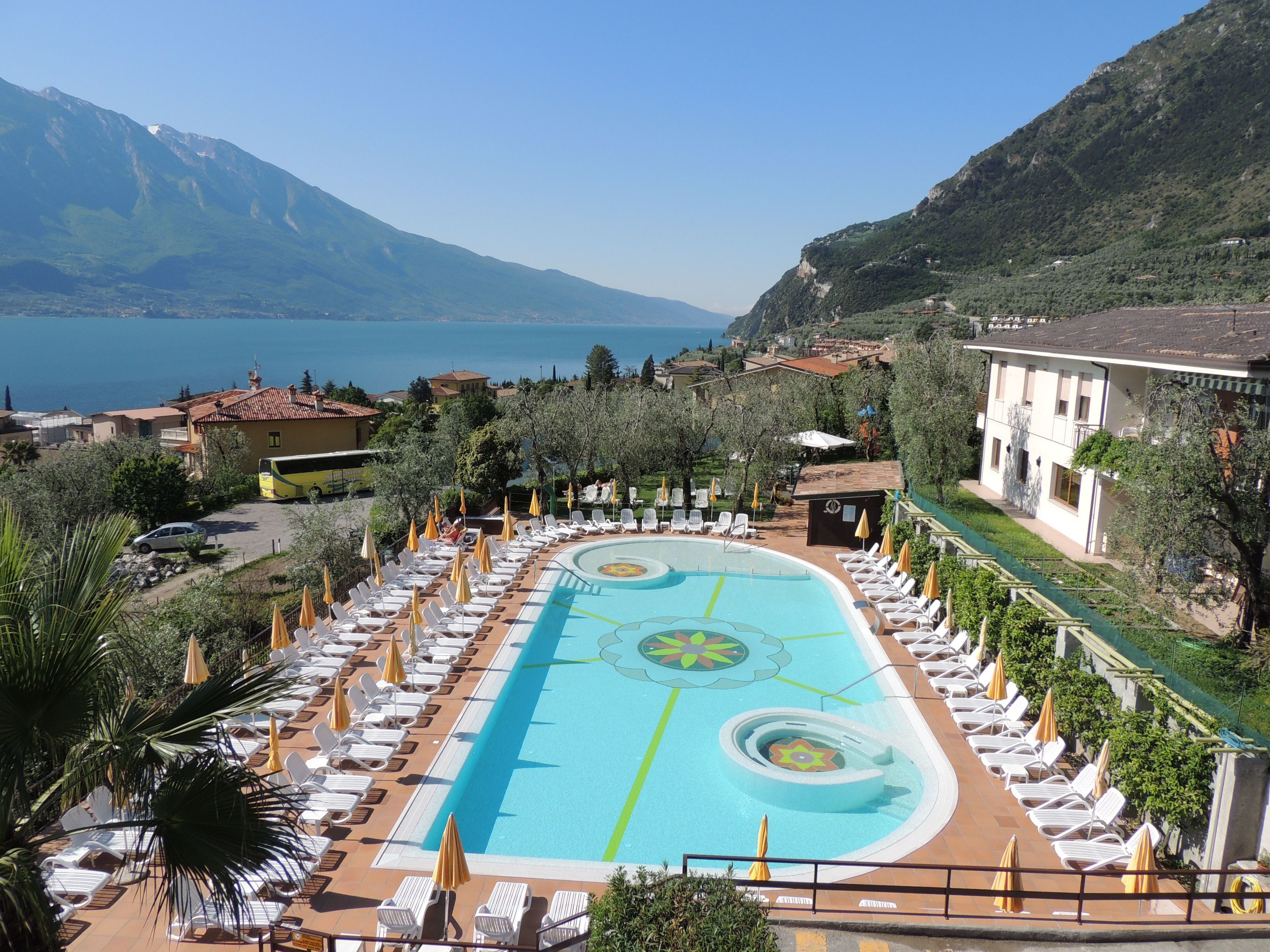 A pool area with lounge chairs and umbrellas, surrounded by greenery and overlooking a body of water and mountains.