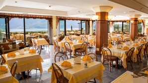 Un restaurante con varias mesas y sillas, amplias ventanas que ofrecen vistas a un cuerpo de agua y montañas, e iluminación interior cálida.