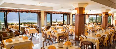 A restaurant with multiple tables and chairs, large windows with a view of a body of water and mountains, and a warm interior lighting.