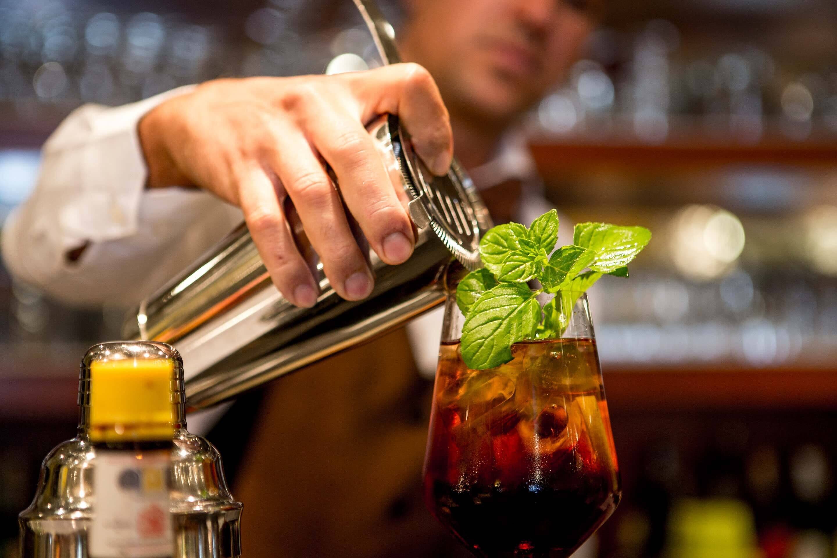 A bartender pouring a drink into a glass with ice and a mint garnish.