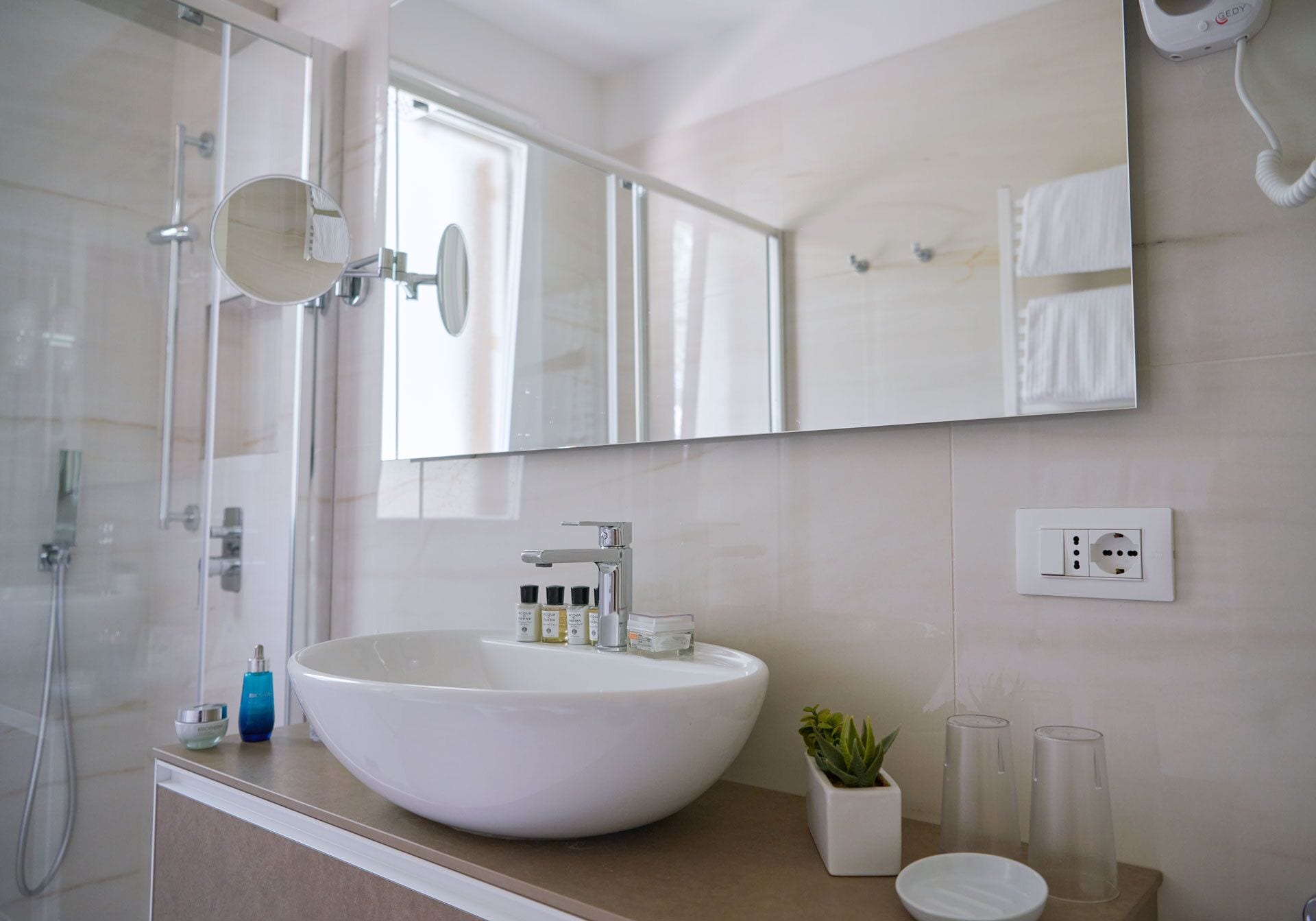 A modern bathroom with a large white sink, a mirror, a shower, and a small potted plant.