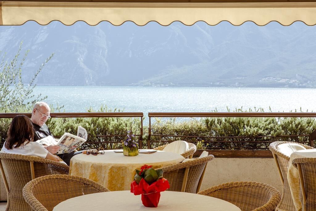 A terrace with wicker chairs and tables, overlooking a lake with mountains in the background.