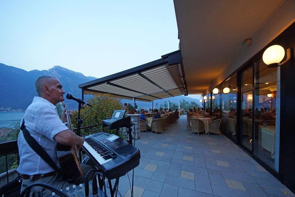 Een man bespeelt een keyboard op een terras in de openlucht, met uitzicht op bergen en een meer.