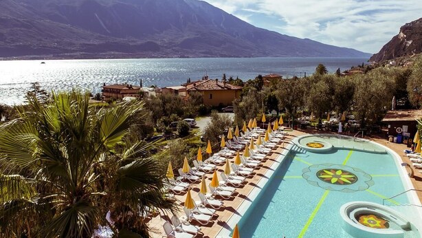 Un área junto a la piscina con sillas de descanso y sombrillas, una piscina con un diseño decorativo y montañas al fondo.