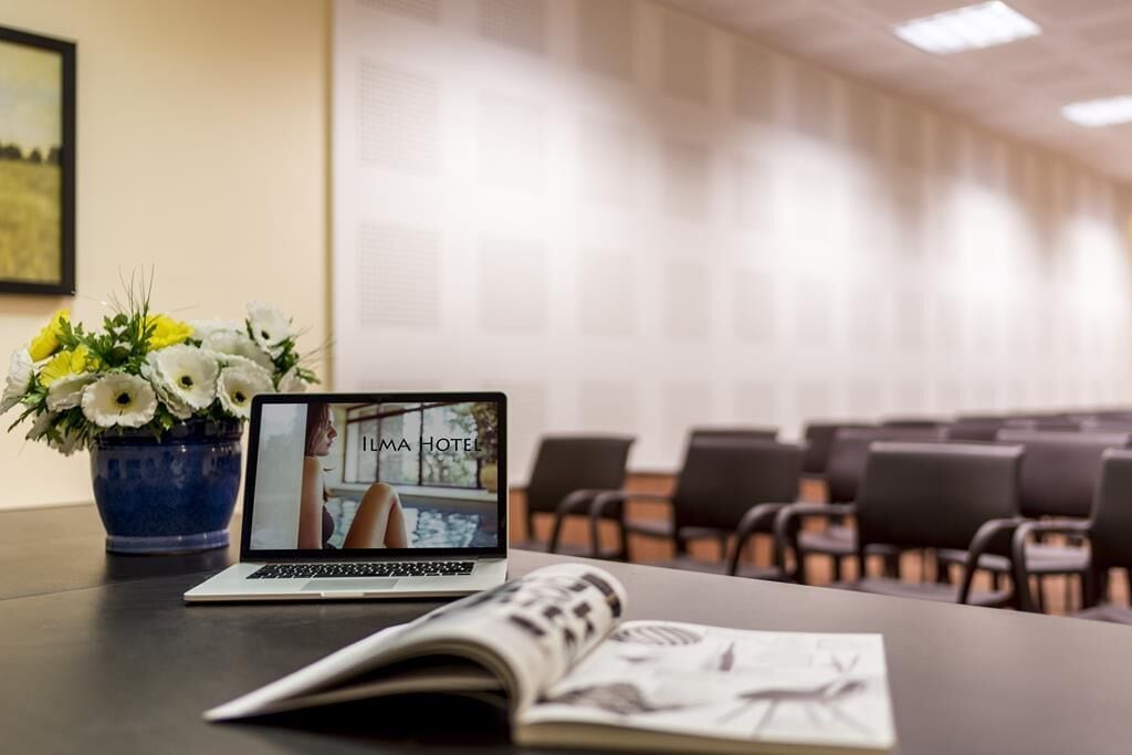 A laptop displaying a hotel interior, a vase with flowers, and a magazine on a table in a conference room.