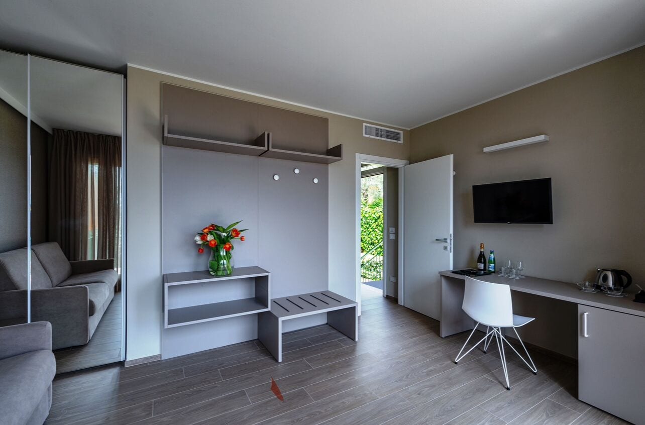 A modern, minimalist living room with a grey sofa, a small bench, and a coffee table. There is a flat-screen TV mounted on the wall, a small desk with a chair, and a vase with flowers on a shelf.