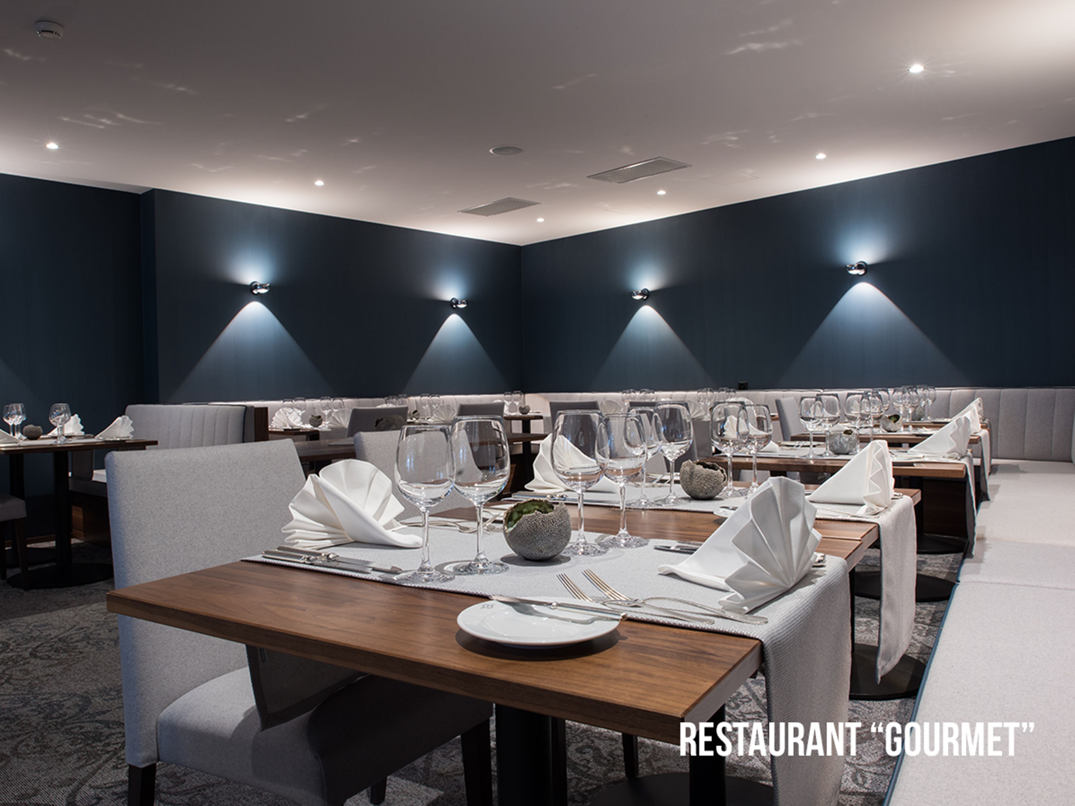 Un restaurant au décor moderne, avec des murs sombres, un éclairage discret et des tables soigneusement disposées, recouvertes de nappes blanches et agrémentées de verres à vin.