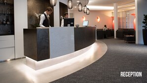A reception area with a curved reception desk, pendant lighting, a seating area with sofas, and a bar with bottles and glasses. - Grand Hôtel Les Endroits (La Chaux-de-Fonds)