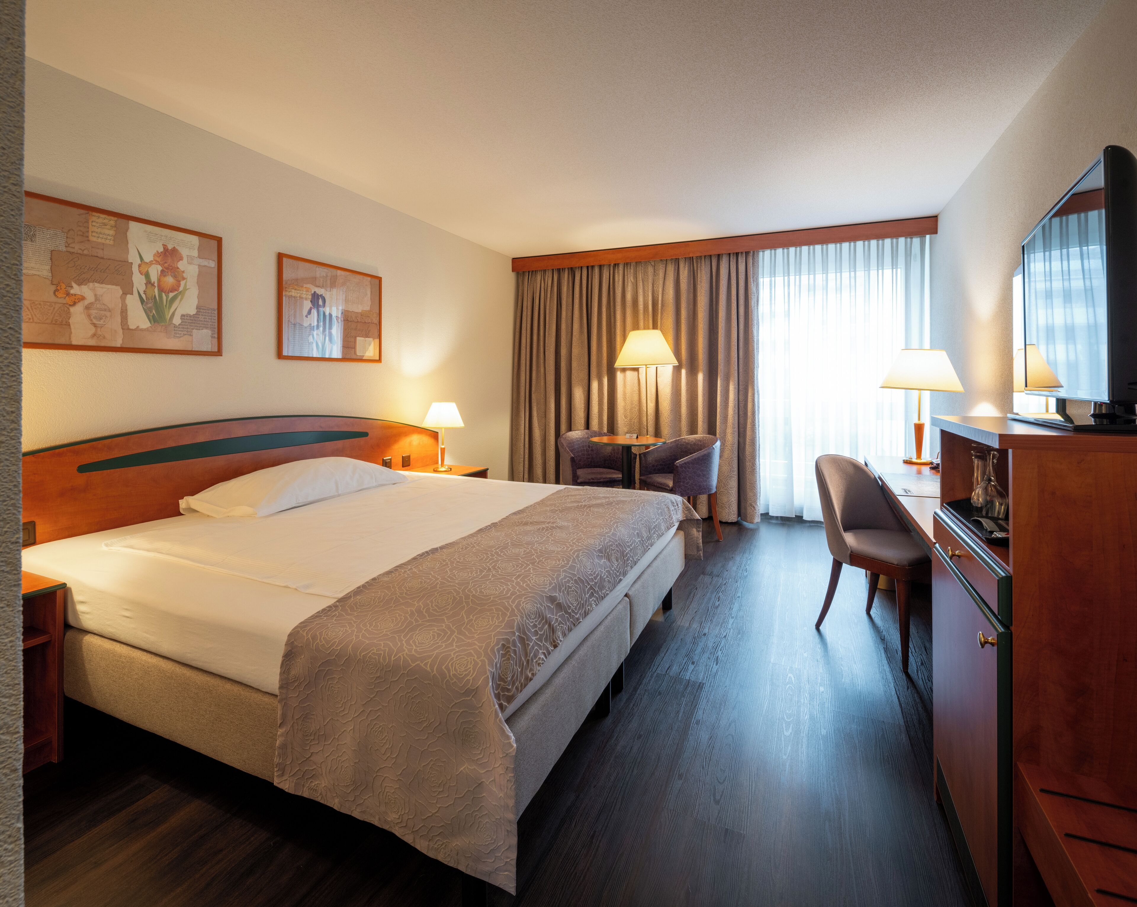A hotel room with a large bed, a desk with a chair, a television, and two lamps.