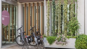 A modern building entrance with two bicycles parked outside, a glass door, and a vertical garden with climbing plants and flowers.