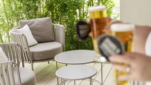 A patio area with wicker furniture and round tables, surrounded by greenery.