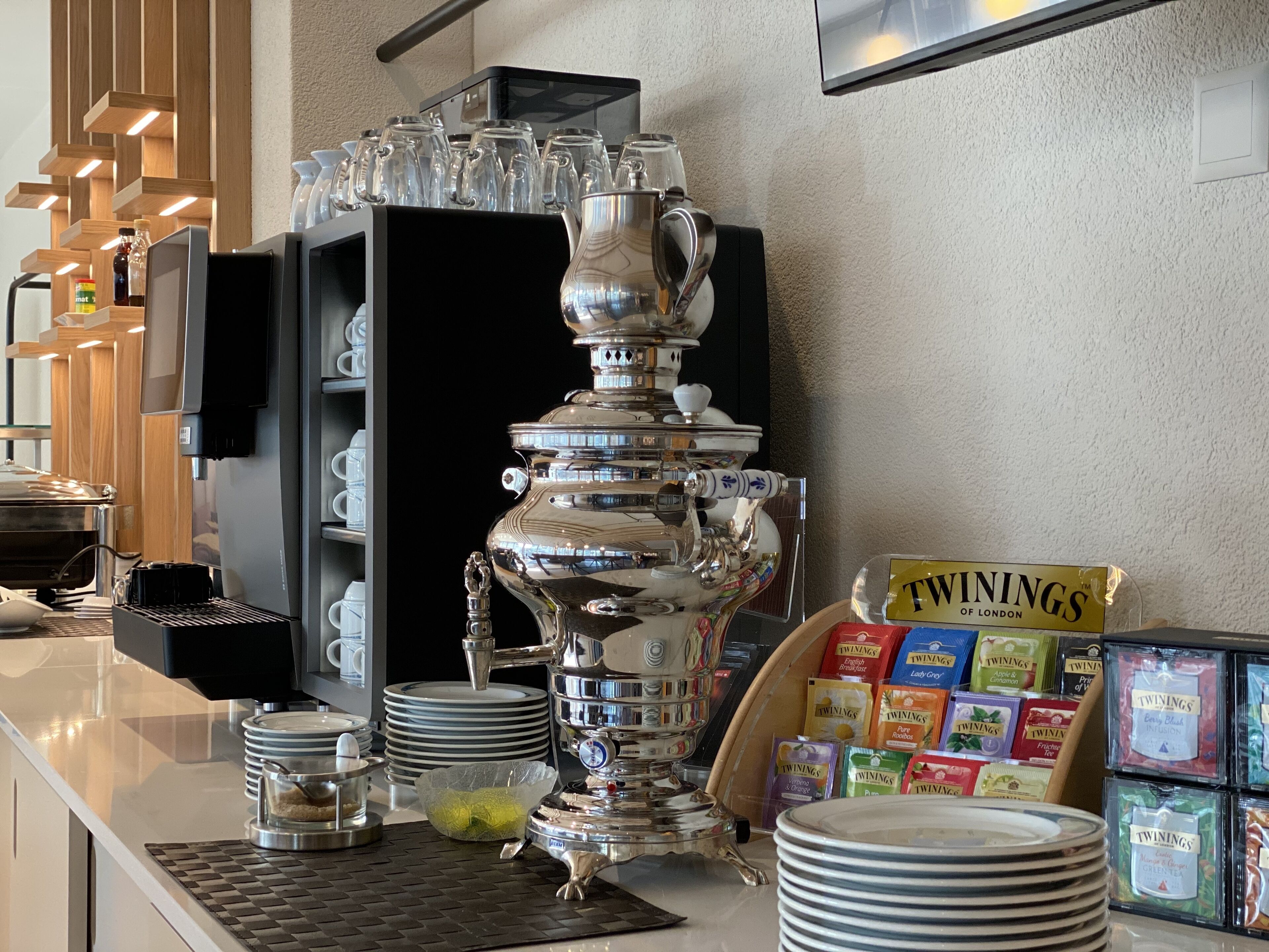 Una estación de café con una cafetera plateada grande, platos apilados y cajas de té Twinings.