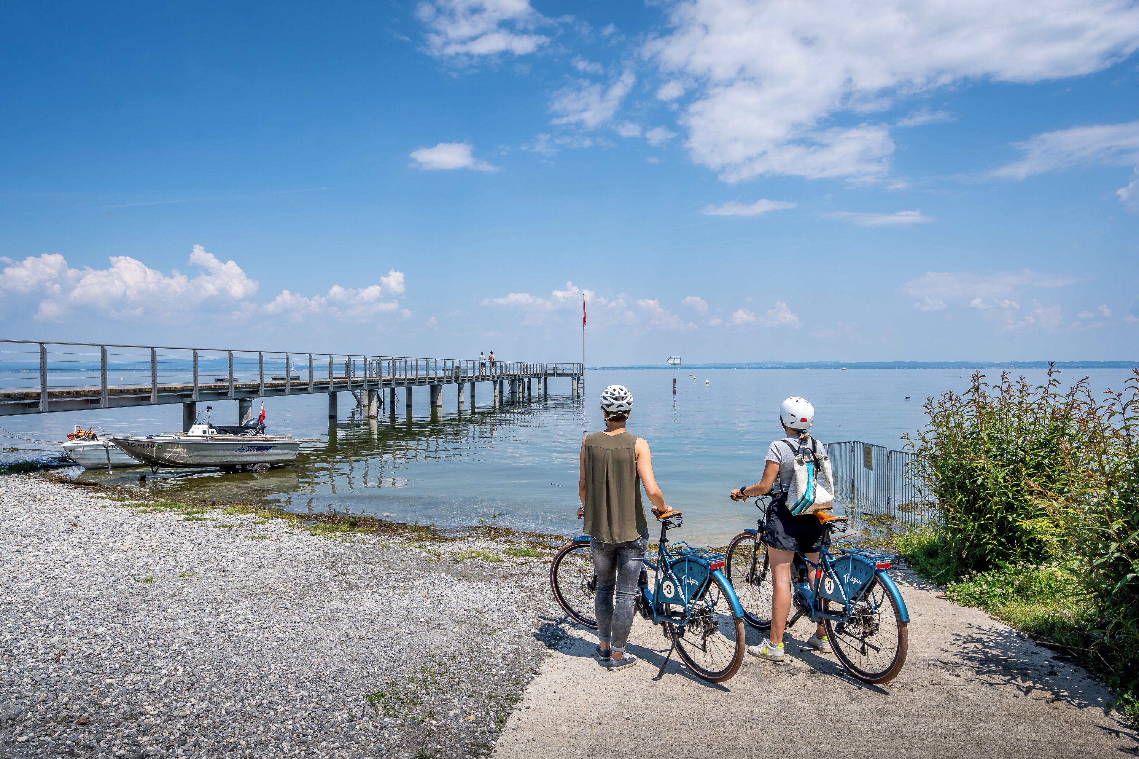 Une scène côtière avec une jetée s’avançant dans des eaux calmes, deux vélos garés sur un sentier et un ciel dégagé.