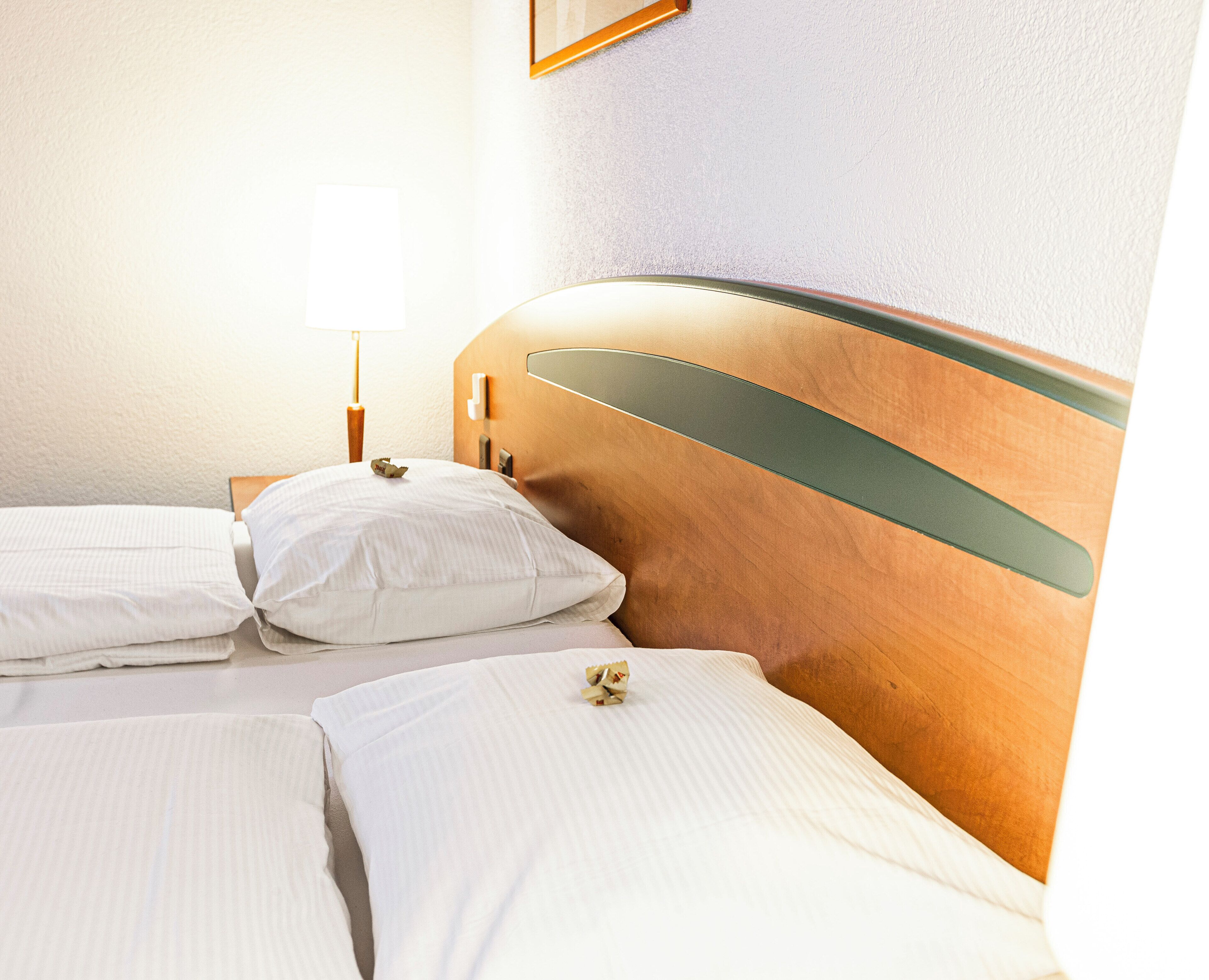 A hotel room with two beds, wooden headboards, white linens, a bedside table with a lamp, and a wall-mounted light fixture.