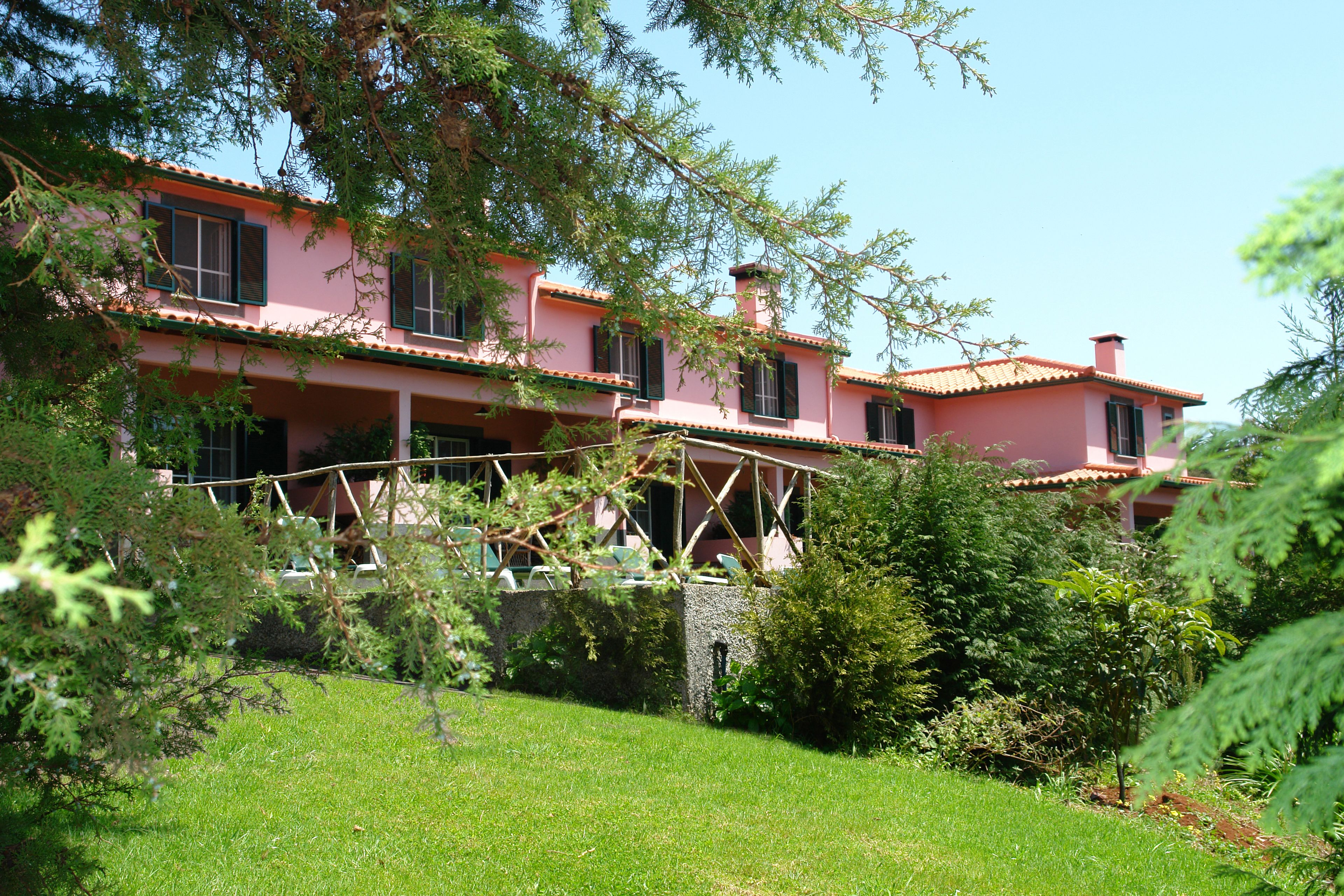 Una villa rosa con techo de tejas rojas, rodeada de exuberante vegetación y un muro de piedra.