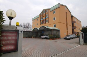 A hotel entrance with a sign indicating the name and number. There are cars parked in front, and the building has a modern design with multiple windows and a flat roof.