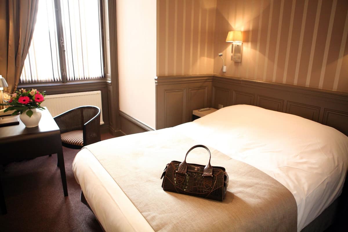 Une chambre d’hôtel avec un lit, un bureau agrémenté d’un vase de fleurs, une chaise et une lampe fixée au mur.