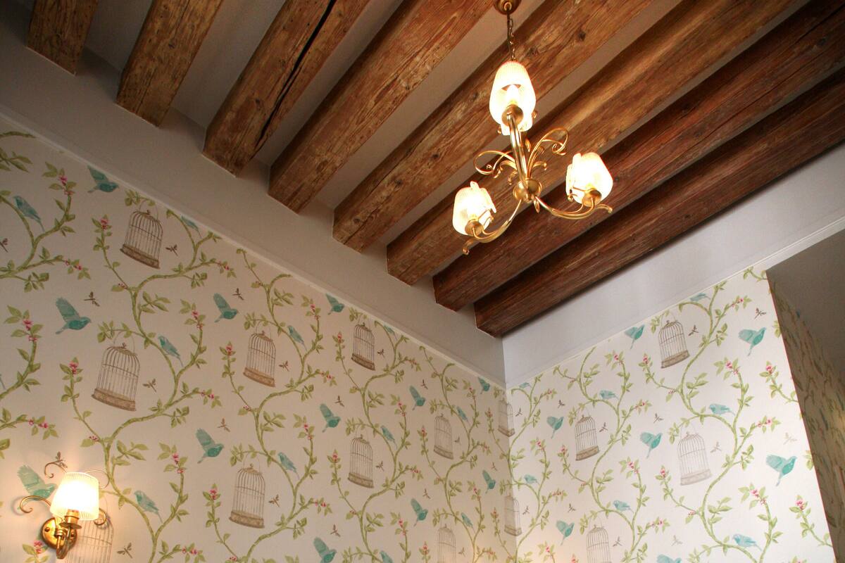 Une chambre avec un plafond en bois, un lustre, un lit avec une tête de lit blanche et un papier peint à motifs floraux.