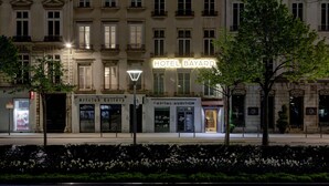 La façade d’un hôtel, sur laquelle on peut voir une enseigne bien visible indiquant « HÔTEL BAYARD », ainsi que les devantures de boutiques nommées « Artclub Gallery » et « Capital Audition », et un trottoir bien entretenu bordé d’arbres.