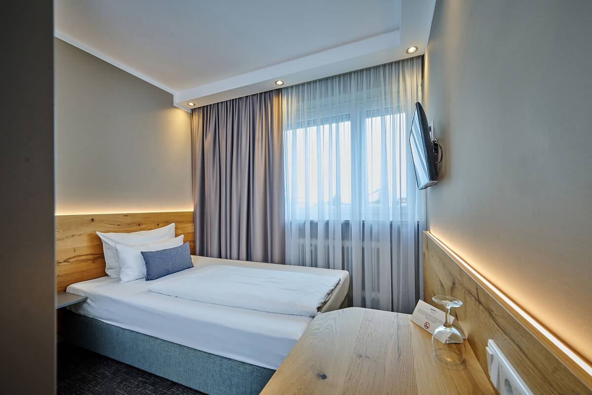 A hotel room with a bed, a bedside table, a TV mounted on the wall, and a window with curtains.