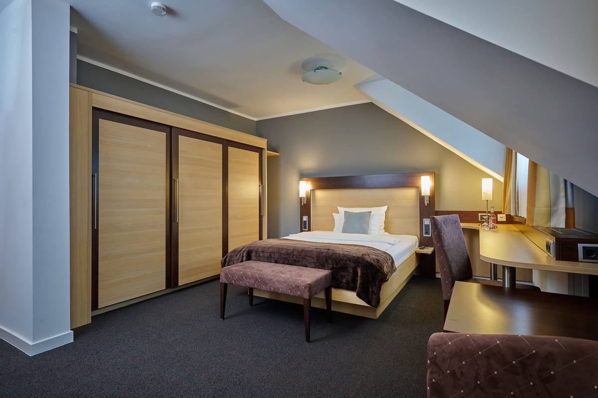 A modern hotel room with a large bed, a desk, a chair, and a wardrobe.