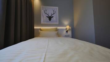 A hotel room with a large bed, a headboard with a light fixture, a wall-mounted picture of a deer head, and a bedside table with a lamp.