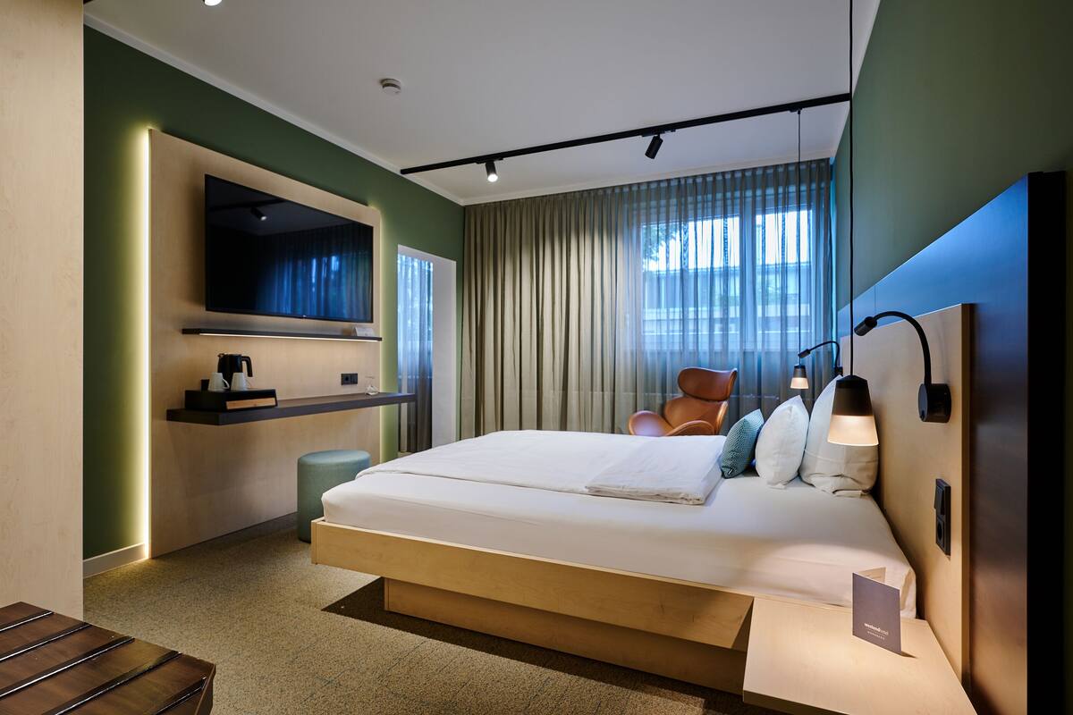 A modern hotel room with a large bed, a flat-screen TV mounted on the wall, a bedside table, and a chair.