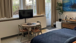 Un dormitorio con cama, escritorio y silla, televisor, planta y un cuadro en la pared.